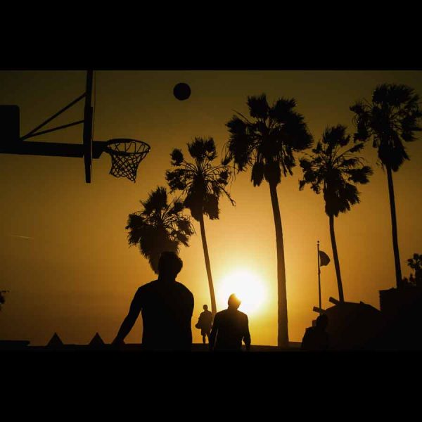 Fotografía de niños jugando en una cancha de baloncesto con el atardecer y palmeras de fondo